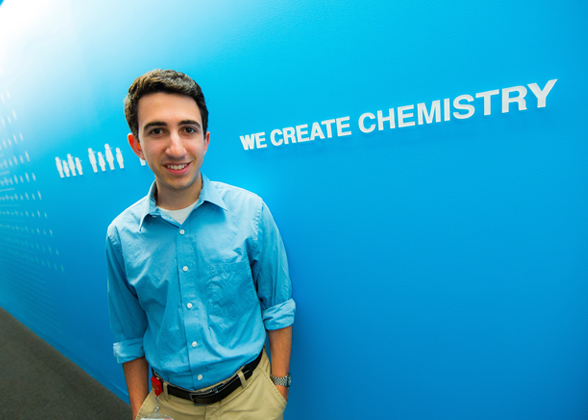 A male student standing in front of a sign that says, "We create chemistry."