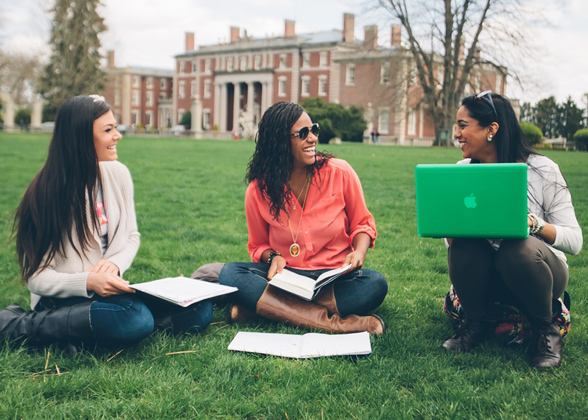 Three students with notebooks and a computer sitting on the lawn outside of Hennessy Hall