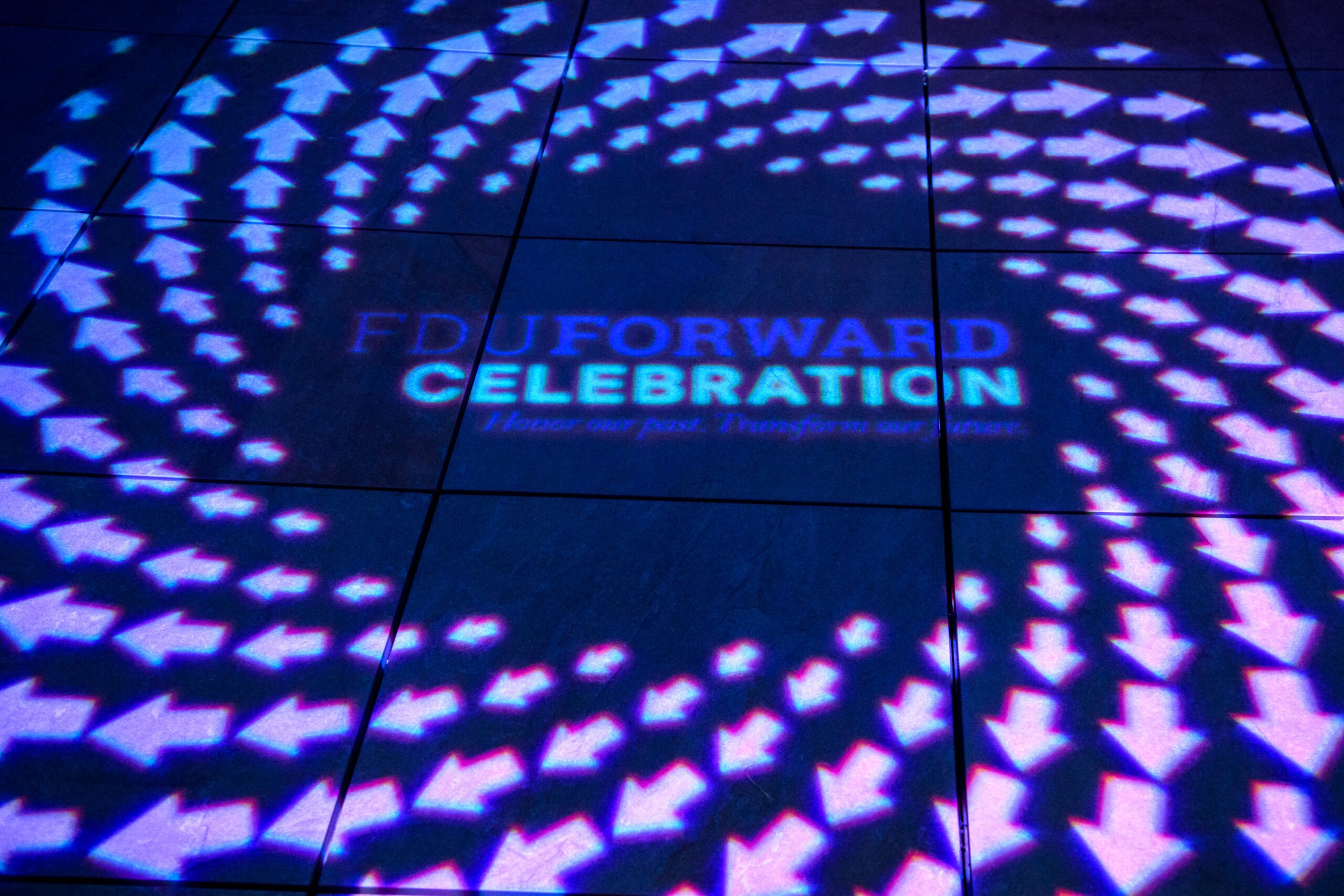 Projected light display on a floor reading “FDU Forward Celebration: Honor our past. Transform our future,” surrounded by circular patterns of illuminated arrows.